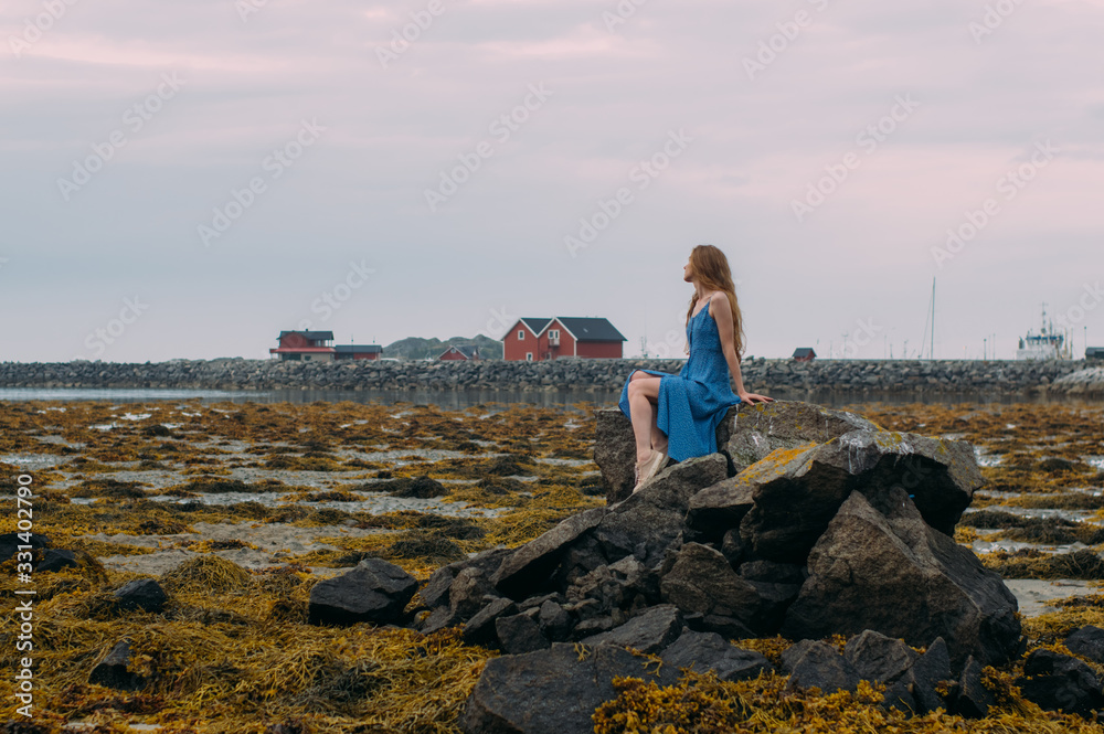 Young woman in blue dress sitting on stone among algae in tide