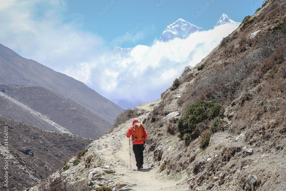 Person in orange windbreaker walks on footpath during sunny day in Himalayas. Kangtega and Thamserku mountain peaks are visible above white clouds. Theme of trekking in Nepal.