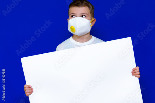 Portrait of caucasian boy in a protective mask respirator holds a blank sheet of paper on a bright blue isolated background. Protection against coronavirus.Mock up,copy space,advertisement. photo