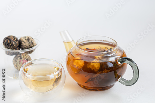 Blooming black tea in a glass teapot. On a light surface. Nearby are tea briquettes of leaves and flowers. In the background is a cake.
