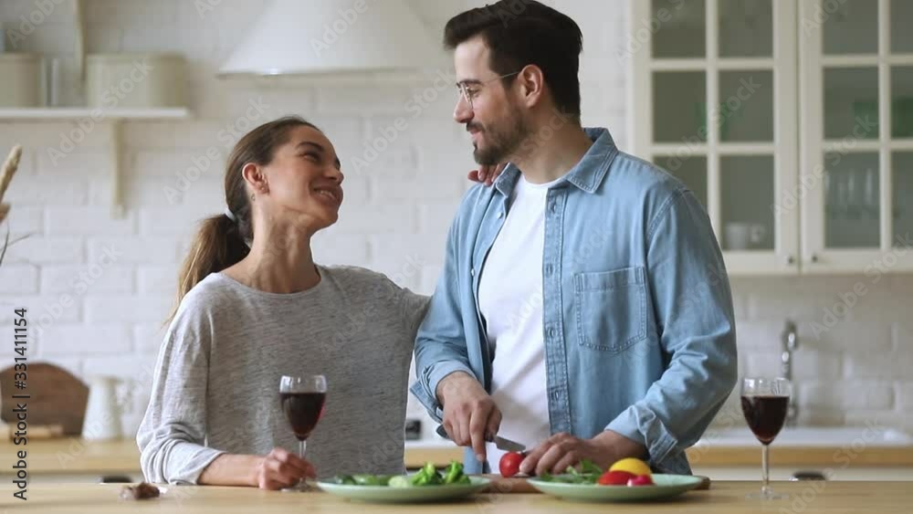 Happy mixed race young woman holding glass of wine, touching shoulder of smiling husband in eyewear chopping vegetables for romantic dinner. Head shot family couple enjoying preparing food together. Happy mixed race young woman holding glass of wine, touching shoulder of smiling husband in eyewear chopping vegetables for romantic dinner. Head shot family couple enjoying preparing food together.