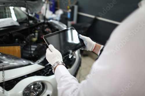 Close-up view of mechanic looking on car through digital tablet with black screen and copy space. Open trunk of white automobile. Auto service station concept