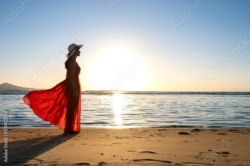 Young woman wearing long red dress and straw hat standing on sand beach at sea shore enjoying view of rising sun in early summer morning.