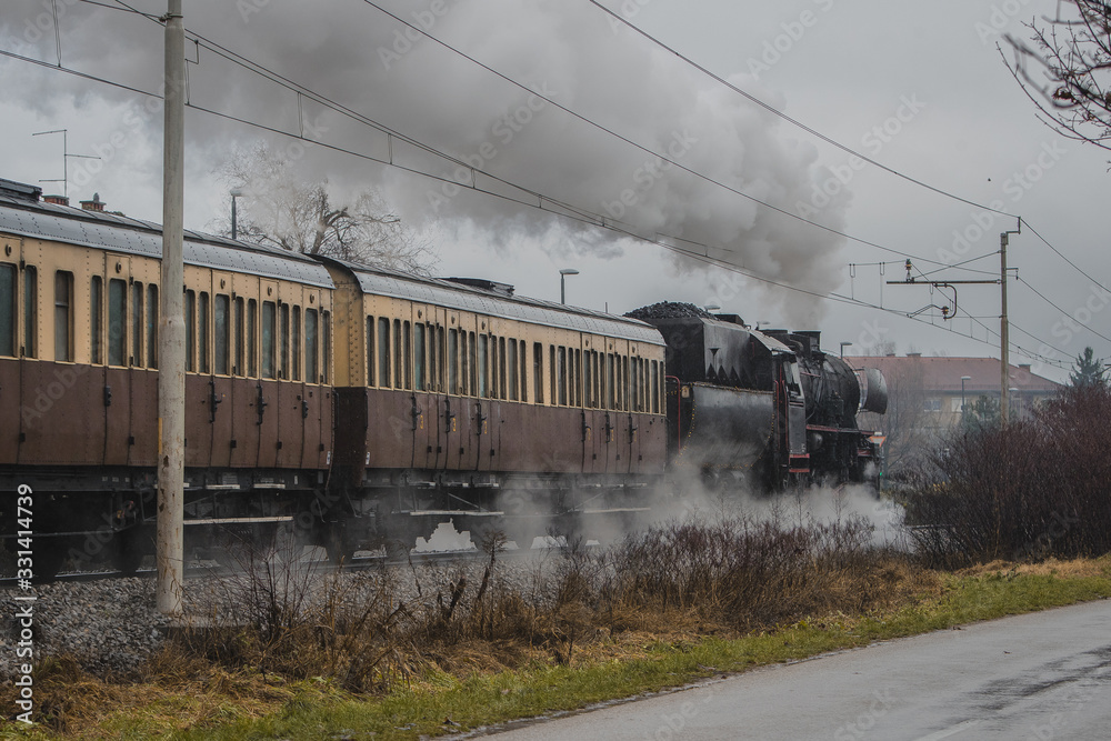 Old vintage steam train with black locomotive is running full power on a railroad track in the city during cold rainy season.