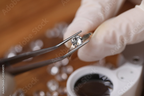 Close-up view of persons hands in protective gloves holding special equipment and examining quality of diamond. Magnifying loupe on table. Luxury jewelry concept