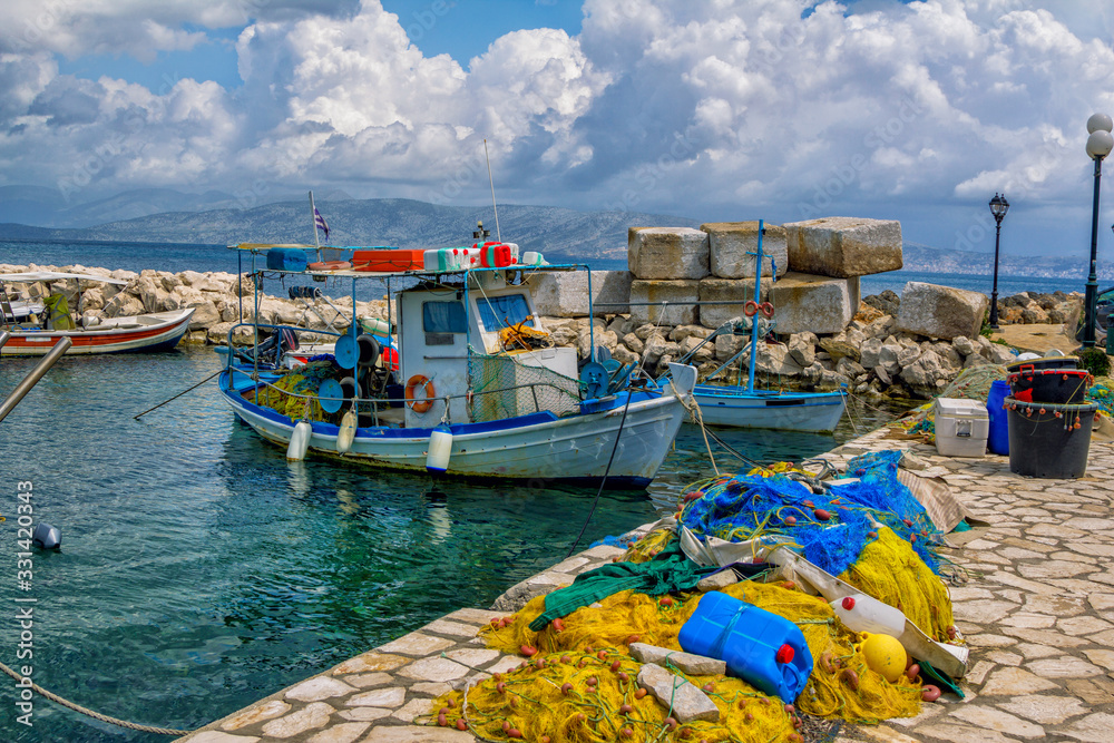 Beautiful landscape with old small fisher boat, colorful fishing nets, calm sea water, blue sky with clouds and mountains on the horizon. Corfu Island, Greece.