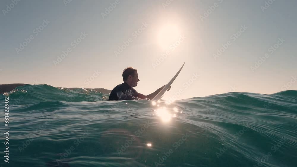 Shot from the water of a male surfer sitting on surfboard in the blue ocean waiting for next big wave. The sun is shining. Surfer easy lifestyle. Extreme sport and meditation.