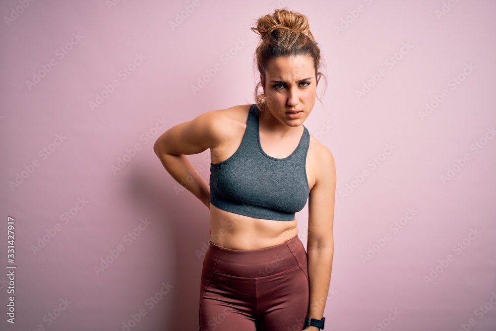 Young beautiful blonde sportswoman doing sport wearing sportswear over pink background Suffering of backache, touching back with hand, muscular pain