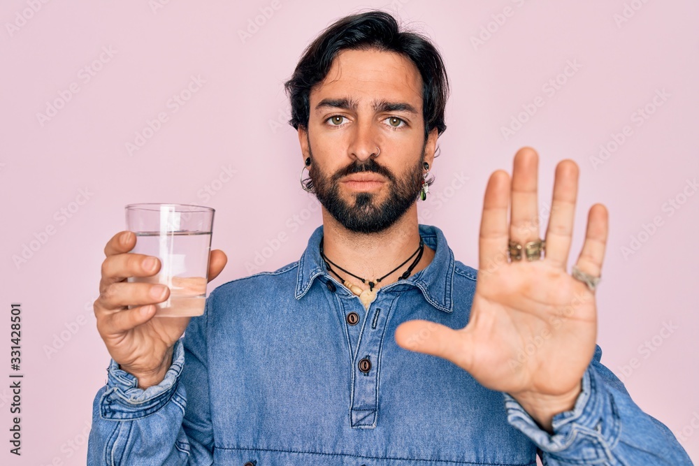 Young handsome hispanic bohemian hipster man with bear drinking a fresh glass of water with open hand doing stop sign with serious and confident expression, defense gesture Young handsome hispanic bohemian hipster man with bear drinking a fresh glass of water with open hand doing stop sign with serious and confident expression, defense gesture