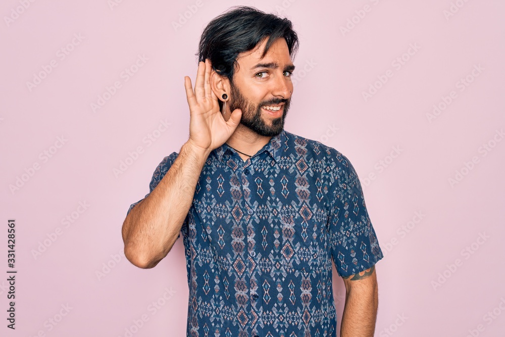 Young handsome hispanic bohemian man wearing hippie style over pink background smiling with hand over ear listening an hearing to rumor or gossip. Deafness concept. Young handsome hispanic bohemian man wearing hippie style over pink background smiling with hand over ear listening an hearing to rumor or gossip. Deafness concept.