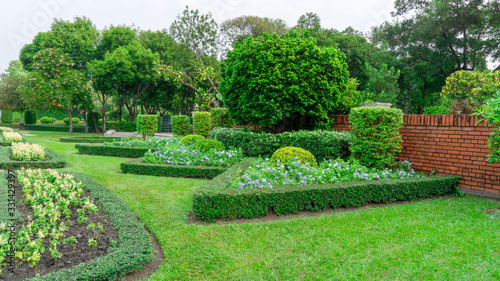 Gardens with geometric shape of bush, decoration flowering plant blooming, green leaf of Philippine tea plant, greenery trees on background under cloudy sky, in a good care landscapes of a public park