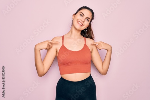 Young beautiful fitness woman wearing sport excersie clothes over pink background looking confident with smile on face  pointing oneself with fingers proud and happy.