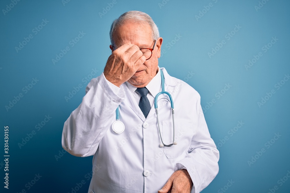 Senior grey haired doctor man wearing stethoscope and medical coat over blue background tired rubbing nose and eyes feeling fatigue and headache. Stress and frustration concept. Senior grey haired doctor man wearing stethoscope and medical coat over blue background tired rubbing nose and eyes feeling fatigue and headache. Stress and frustration concept.