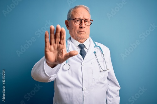 Senior grey haired doctor man wearing stethoscope and medical coat over blue background doing stop sing with palm of the hand. Warning expression with negative and serious gesture on the face.