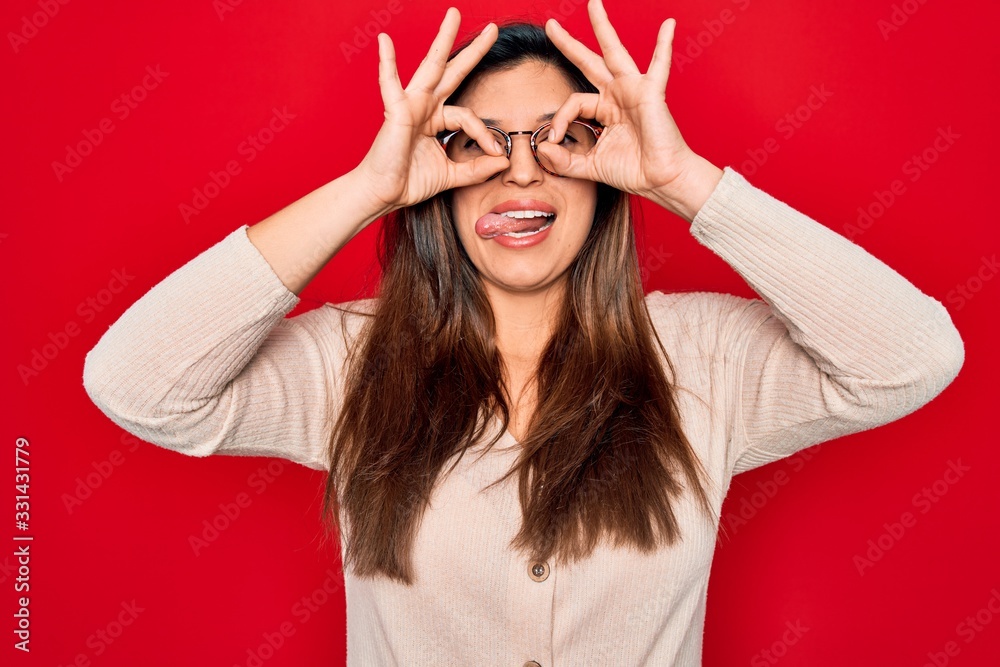 Young hispanic smart woman wearing glasses standing over red isolated background doing ok gesture like binoculars sticking tongue out, eyes looking through fingers. Crazy expression. Young hispanic smart woman wearing glasses standing over red isolated background doing ok gesture like binoculars sticking tongue out, eyes looking through fingers. Crazy expression.