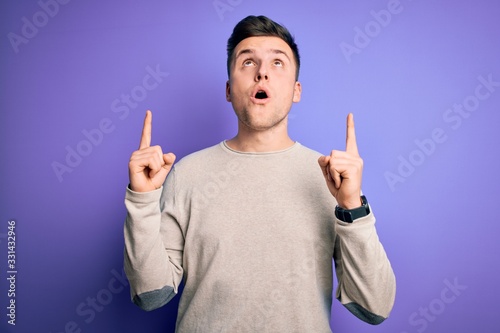 Young handsome caucasian man wearing casual sweater over purple isolated background amazed and surprised looking up and pointing with fingers and raised arms.