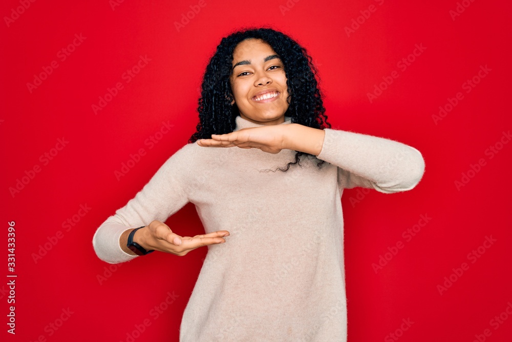 Fototapeta premium Young african american curly woman wearing casual turtleneck sweater over red background gesturing with hands showing big and large size sign, measure symbol. Smiling looking at the camera. Measuring