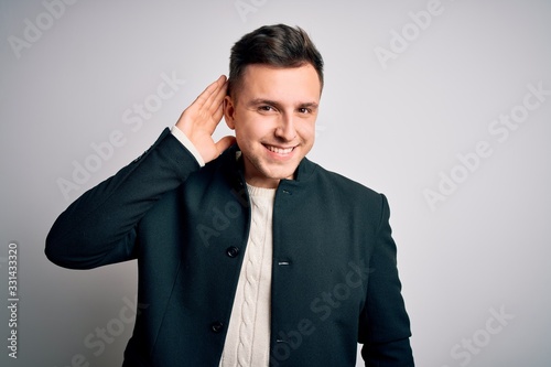 Young handsome business mas wearing elegant winter coat standing over isolated background smiling with hand over ear listening an hearing to rumor or gossip. Deafness concept.