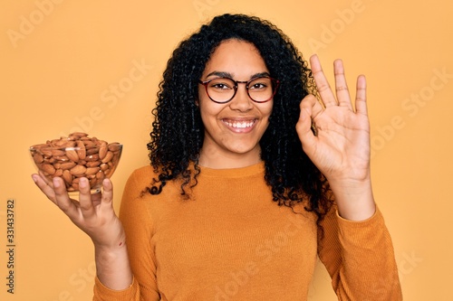 Young african american curly woman holding bowl with healthy almonds over yellow background doing ok sign with fingers  excellent symbol