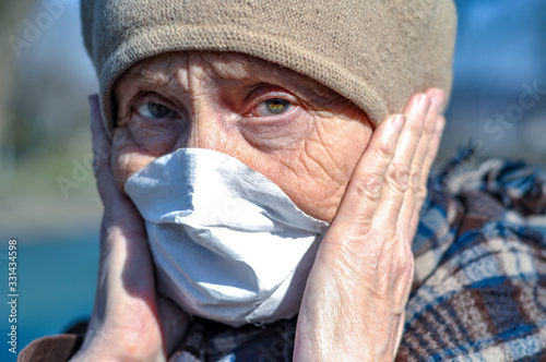 An elderly old woman in a protective respirator mask on her face is terrified holding her hands to her head. Elderly and coronovirus infection COVID-19. Retirement Health at Risk