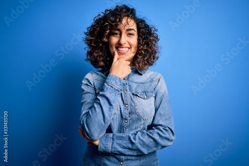 Leinwand Poster Young beautiful curly arab woman wearing casual denim shirt standing over blue background looking confident at the camera smiling with crossed arms and hand raised on chin