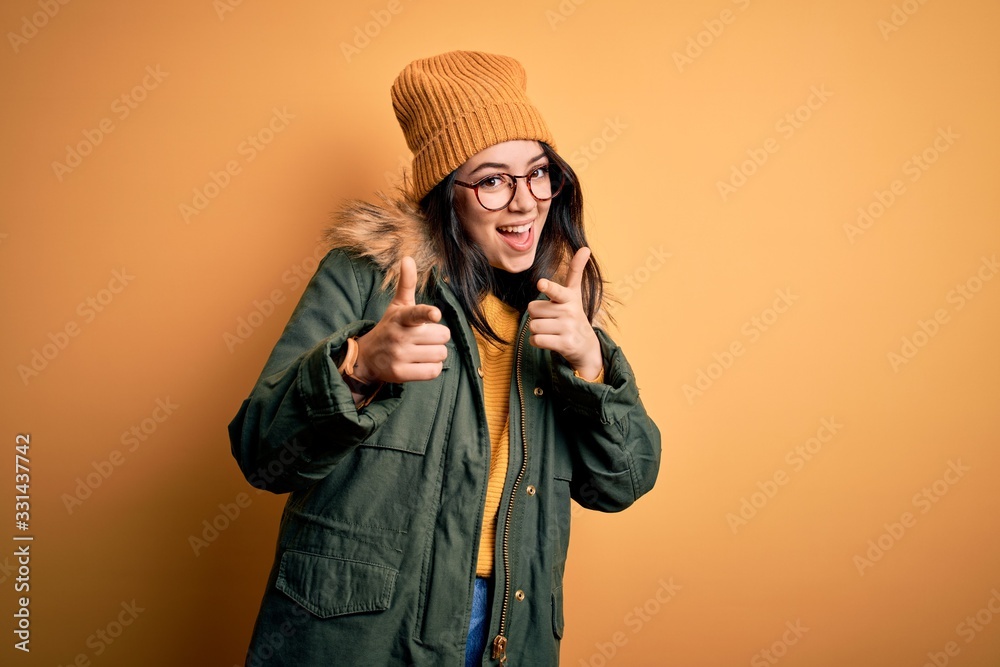 Young brunette woman wearing glasses and winter coat with hat over yellow isolated background pointing fingers to camera with happy and funny face. Good energy and vibes. Young brunette woman wearing glasses and winter coat with hat over yellow isolated background pointing fingers to camera with happy and funny face. Good energy and vibes.