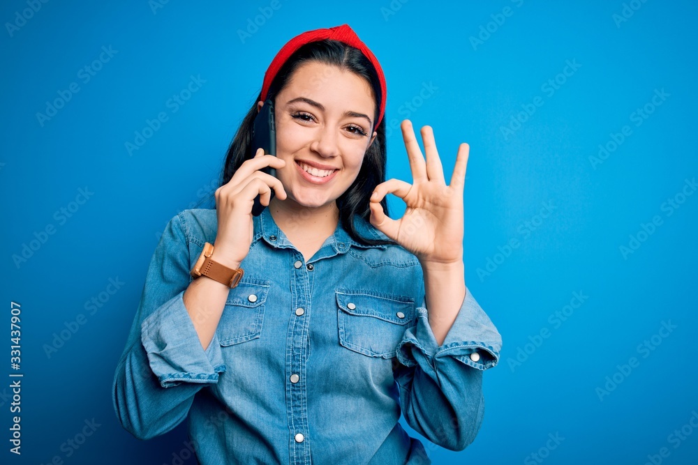 Young brunette woman talking on smartphone over blue isolated background doing ok sign with fingers, excellent symbol Young brunette woman talking on smartphone over blue isolated background doing ok sign with fingers, excellent symbol
