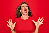 Middle age senior brunette woman wearing casual t-shirt standing over red background crazy and mad shouting and yelling with aggressive expression and arms raised. Frustration concept.