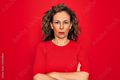 Middle age senior brunette woman wearing casual t-shirt standing over red background skeptic and nervous, disapproving expression on face with crossed arms. Negative person.