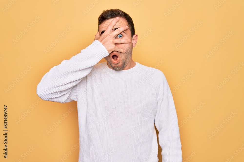 Young handsome man with blue eyes wearing casual sweater standing over yellow background peeking in shock covering face and eyes with hand, looking through fingers with embarrassed expression. Young handsome man with blue eyes wearing casual sweater standing over yellow background peeking in shock covering face and eyes with hand, looking through fingers with embarrassed expression.