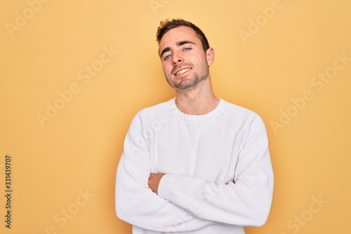 Young handsome man with blue eyes wearing casual sweater standing over yellow background happy face smiling with crossed arms looking at the camera. Positive person.