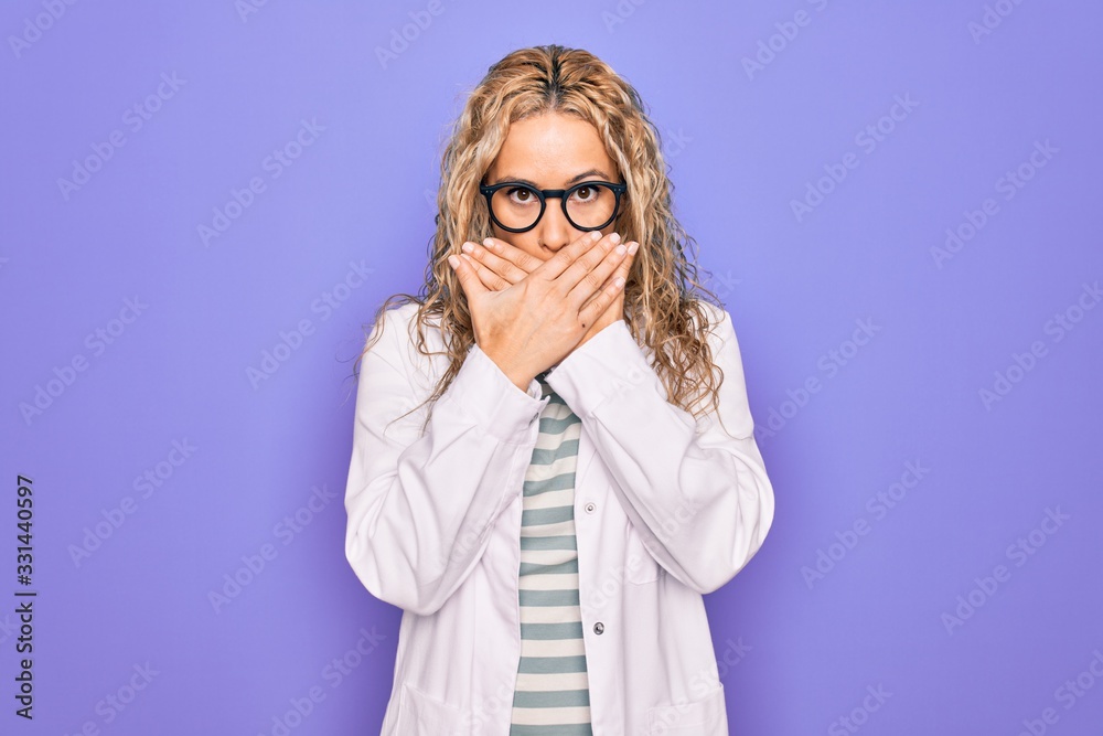 Young beautiful blonde scientist woman wearing coat and glasses over purple background shocked covering mouth with hands for mistake. Secret concept.