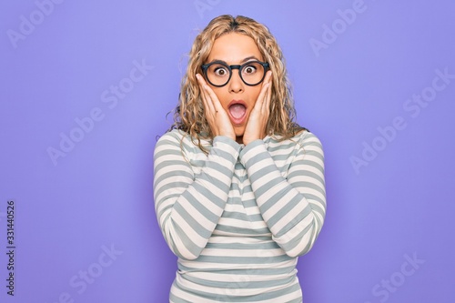 Beautiful blonde woman wearing casual striped t-shirt and glasses over purple background afraid and shocked, surprise and amazed expression with hands on face © Krakenimages.com