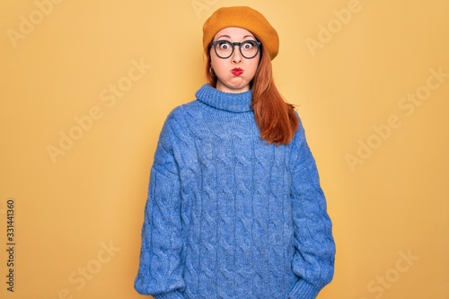 Young beautiful redhead woman wearing french beret and glasses over yellow background puffing cheeks with funny face. Mouth inflated with air, crazy expression. © Krakenimages.com