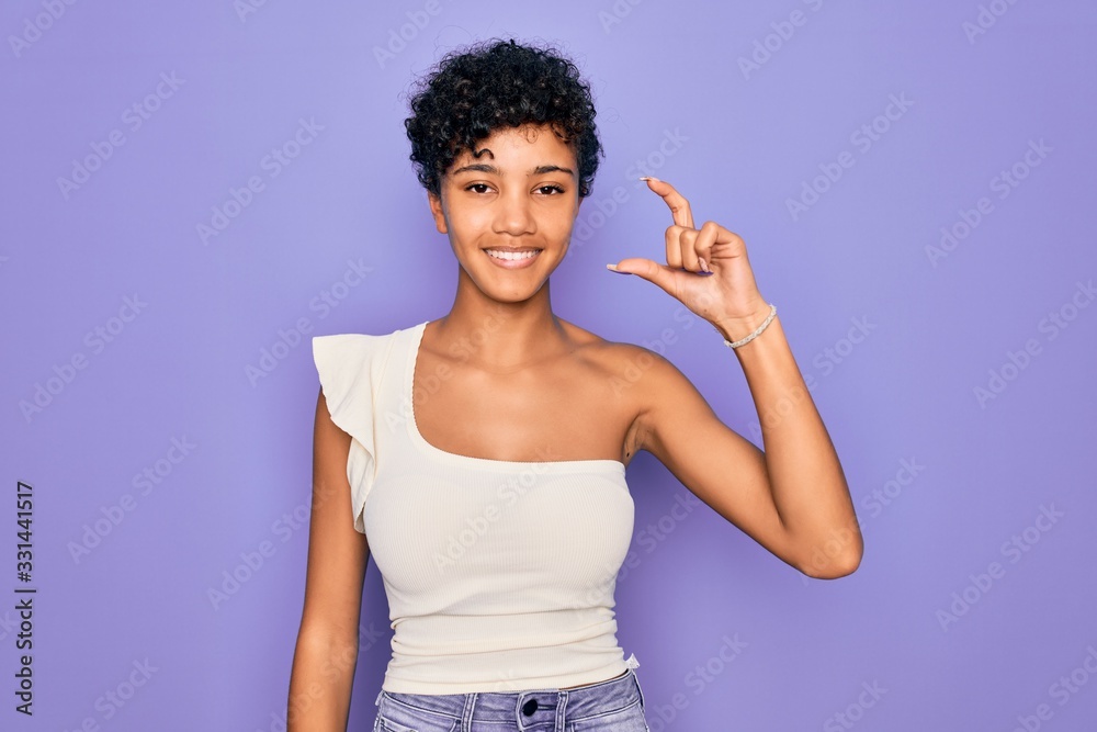 Young beautiful african american afro woman wearing casual t-shirt over purple background smiling and confident gesturing with hand doing small size sign with fingers looking and the camera. Measure Young beautiful african american afro woman wearing casual t-shirt over purple background smiling and confident gesturing with hand doing small size sign with fingers looking and the camera. Measure