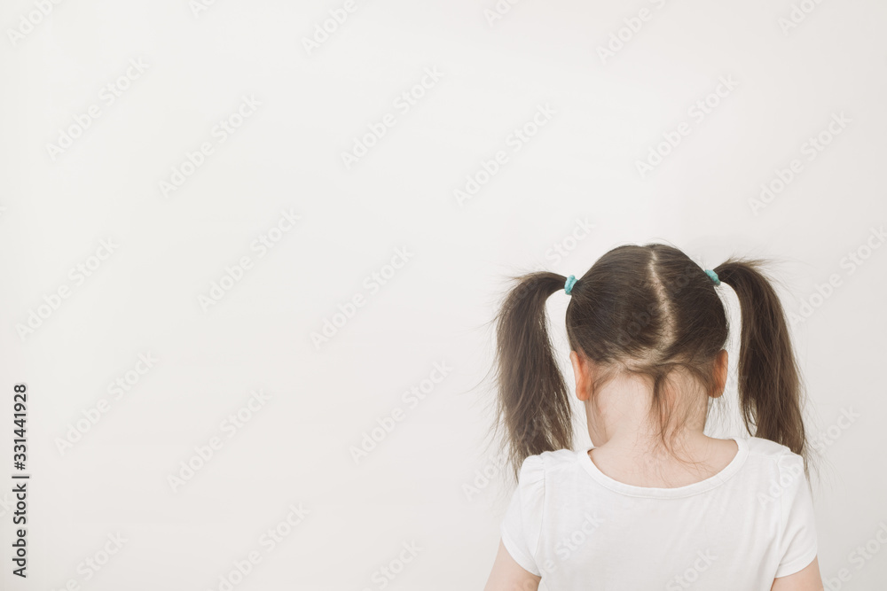 Fototapeta premium A girl with two ponytails stands with her back to the camera. The child turned away to the light wall. Upset baby crying