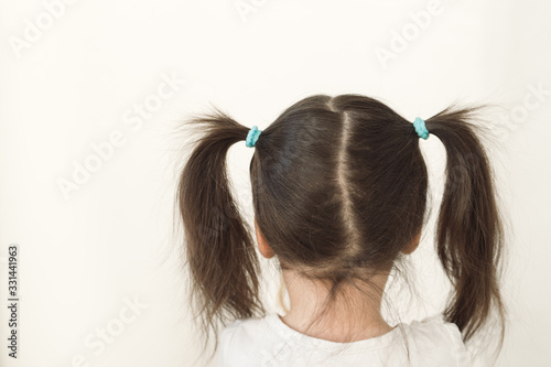 A girl with two ponytails stands with her back to the camera. The child turned away to the light wall. Upset baby crying