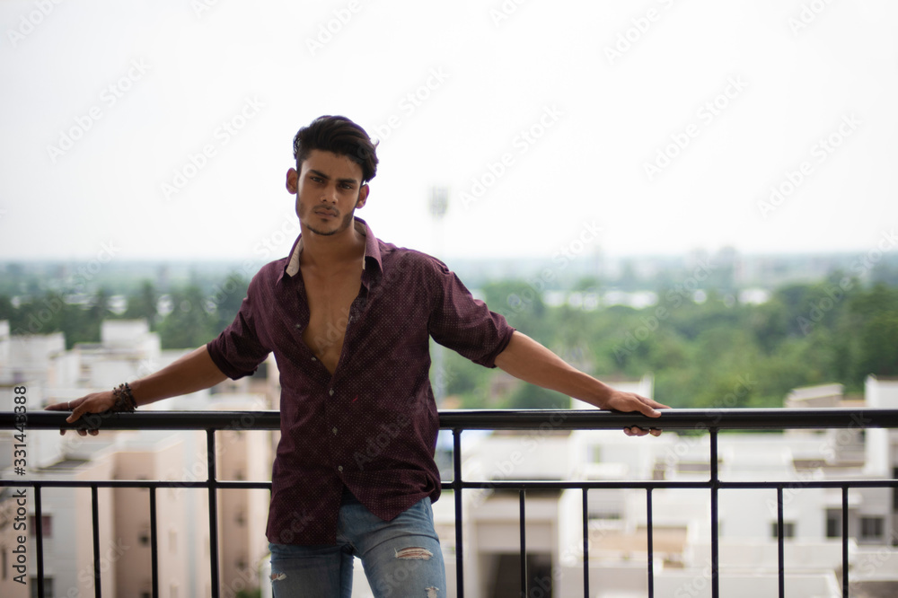 Portrait of an young and handsome brunette Bengali muscular man in casual shirt and jeans standing on a balcony in white urban background. Indian lifestyle.