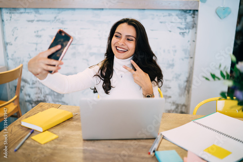 Happy hipster girl using front cellphone camera for clicking selfie pictures during influence vlog conference for communicate with social followers, cheerful freelancer shooting video during mobility photo