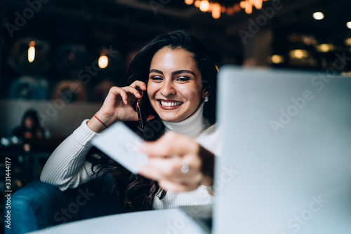 Cheerful female millennial with business card in hand enjoying mobile communication during time for freelance job, successful woman dialing contact number during cellular phoning at table with laptop