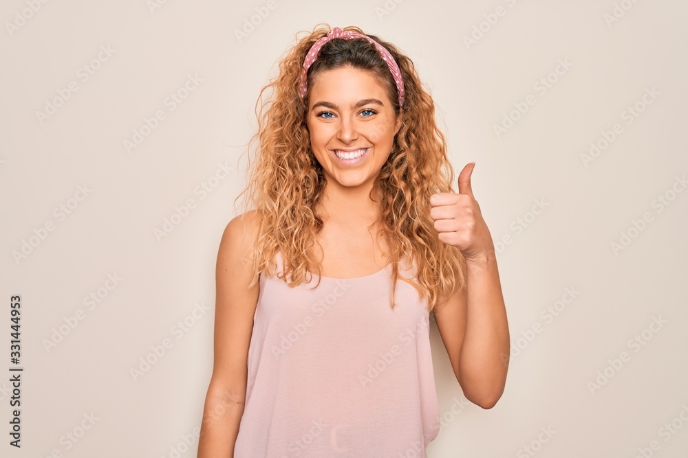 Young beautiful woman with blue eyes wearing casual t-shirt and diadem over pink background doing happy thumbs up gesture with hand. Approving expression looking at the camera showing success. Young beautiful woman with blue eyes wearing casual t-shirt and diadem over pink background doing happy thumbs up gesture with hand. Approving expression looking at the camera showing success.