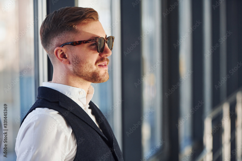 Man in elegant clothes and sunglasses is standing indoors and posing for the camera Man in elegant clothes and sunglasses is standing indoors and posing for the camera