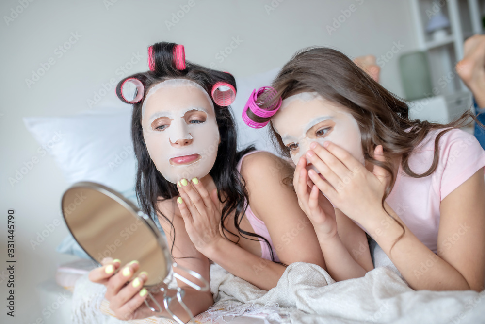 Mom and daughter with facial mask on their faces looking in the mirror Mom and daughter with facial mask on their faces looking in the mirror