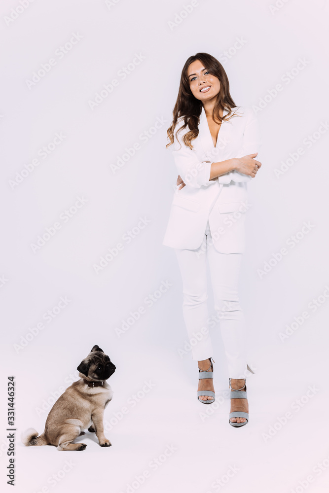 A laughing and smiling auburn haired woman in a white dress and white shoes, is staring most lovingly at her cute pug, who calmly sits down, gaining her undivided attention. Isolated white background.