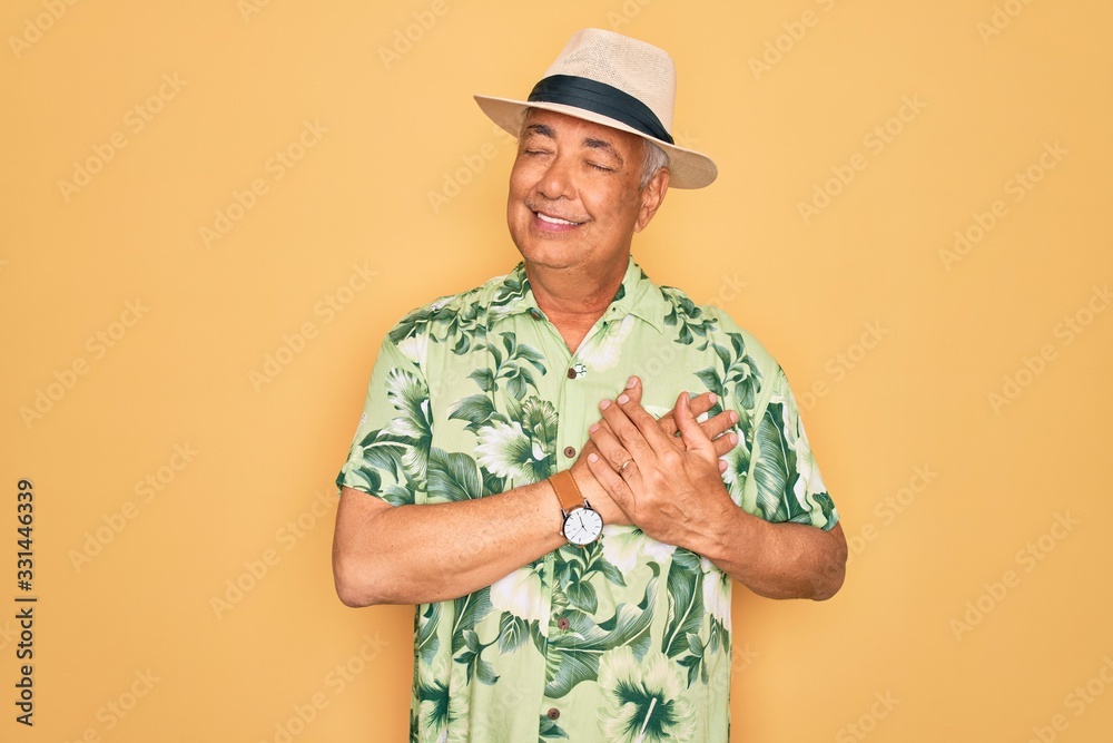 Middle age senior grey-haired man wearing summer hat and floral shirt on beach vacation smiling with hands on chest with closed eyes and grateful gesture on face. Health concept. Middle age senior grey-haired man wearing summer hat and floral shirt on beach vacation smiling with hands on chest with closed eyes and grateful gesture on face. Health concept.