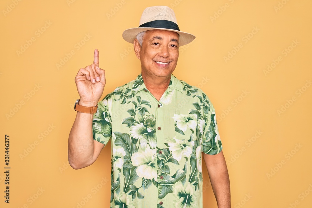 Middle age senior grey-haired man wearing summer hat and floral shirt on beach vacation showing and pointing up with finger number one while smiling confident and happy. Middle age senior grey-haired man wearing summer hat and floral shirt on beach vacation showing and pointing up with finger number one while smiling confident and happy.