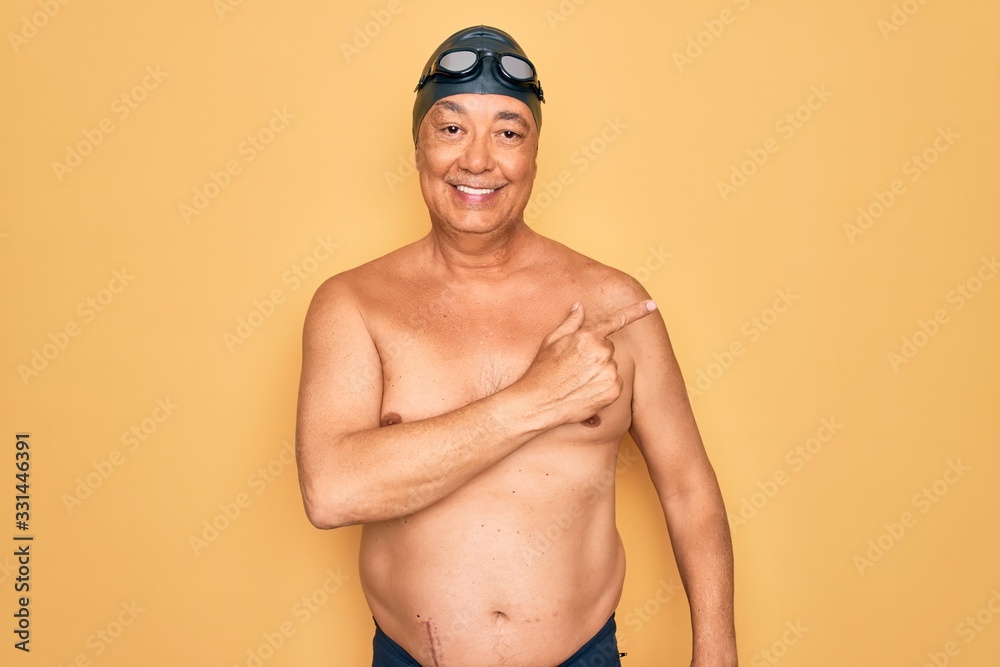 Middle age senior grey-haired swimmer man wearing swimsuit, cap and goggles cheerful with a smile on face pointing with hand and finger up to the side with happy and natural expression