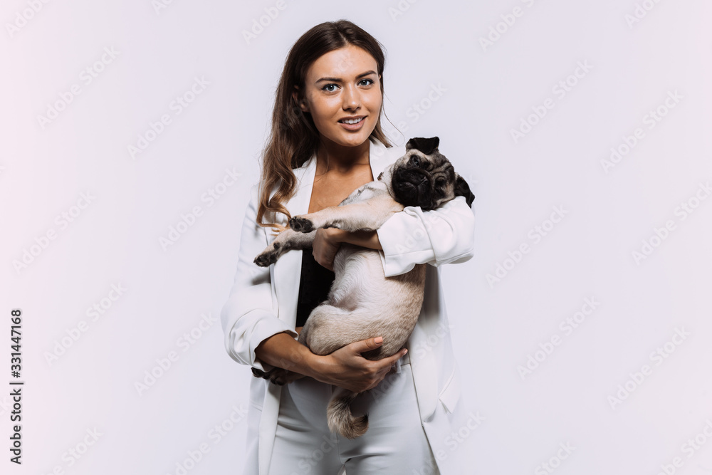 A laughing and smiling auburn haired woman in a white dress, is staring most lovingly at her cute pug, who calmly sits on the hands, gaining her undivided attention. Isolated white background. A laughing and smiling auburn haired woman in a white dress, is staring most lovingly at her cute pug, who calmly sits on the hands, gaining her undivided attention. Isolated white background.