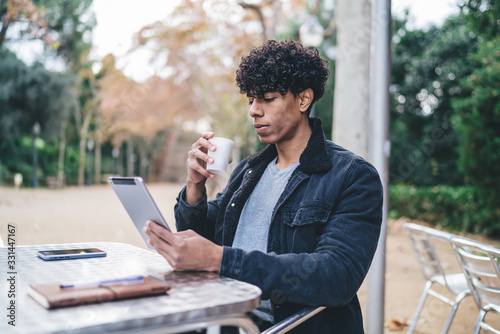 Freelance adult curly male siting at table browsing tablet while drinking coffee in cafe on street