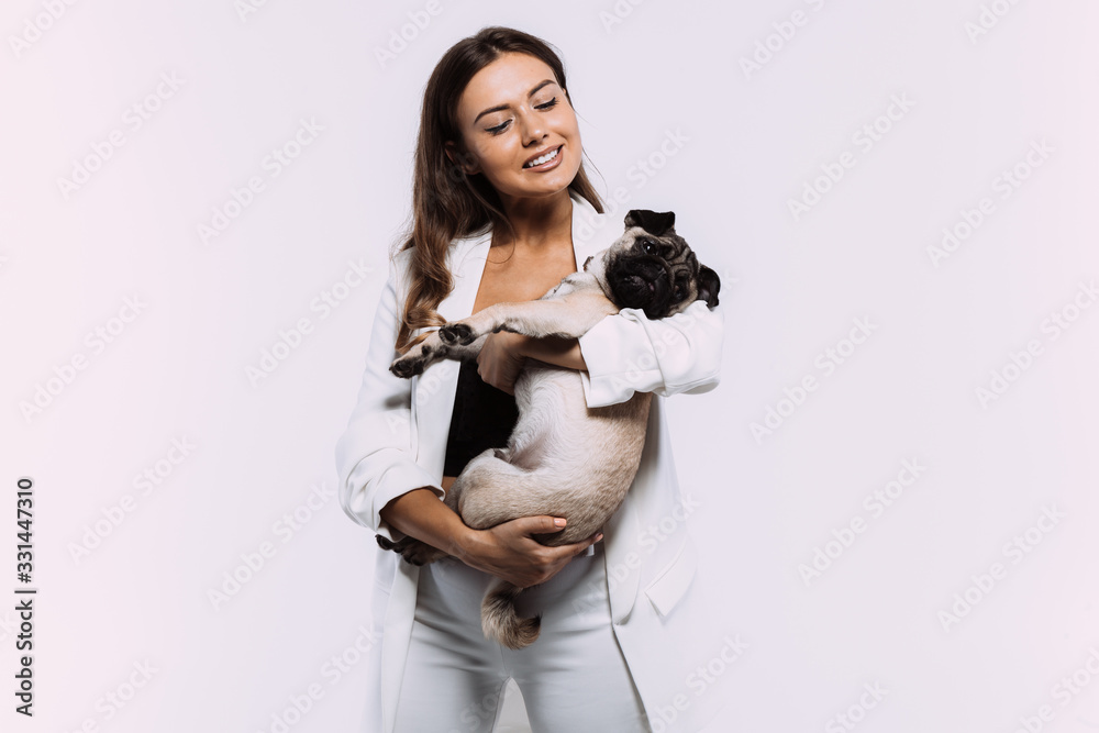A laughing and smiling auburn haired woman in a white dress, is staring most lovingly at her cute pug, who calmly sits on the hands, gaining her undivided attention. Isolated white background. A laughing and smiling auburn haired woman in a white dress, is staring most lovingly at her cute pug, who calmly sits on the hands, gaining her undivided attention. Isolated white background.